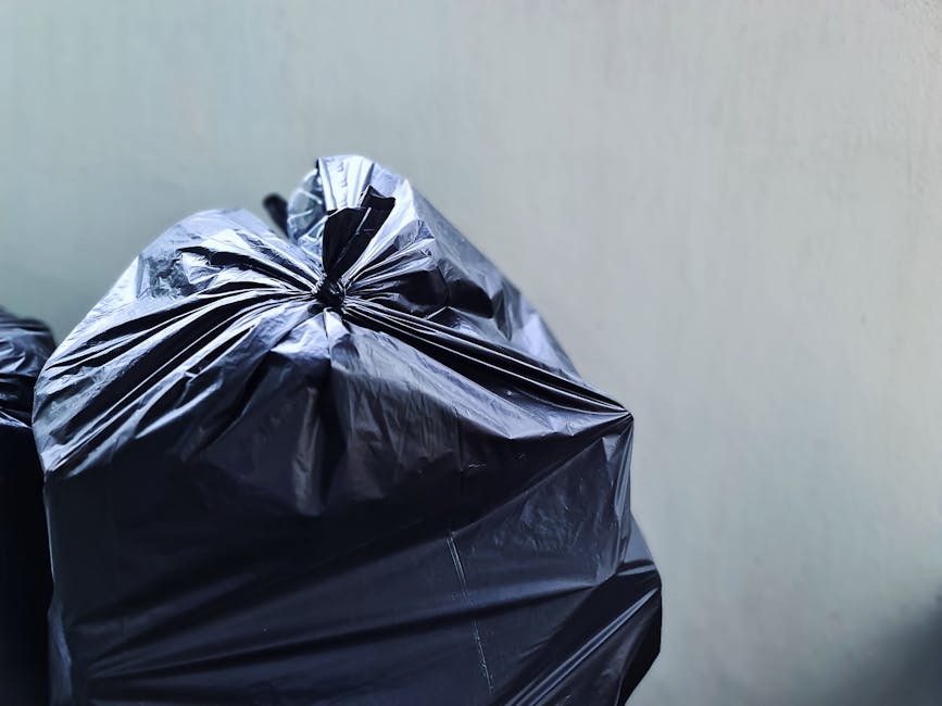 Two large black plastic rubbish bags filled with waste are placed on the pavement beside a wooden fence and a small section of curb. The bags appear to be tightly knotted at the top, with one slightly